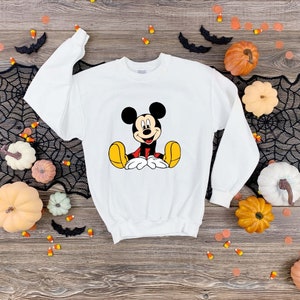 May include: White sweatshirt with a cartoon image of Mickey Mouse sitting down. The sweatshirt is on a wooden surface with pumpkins and candy corn.