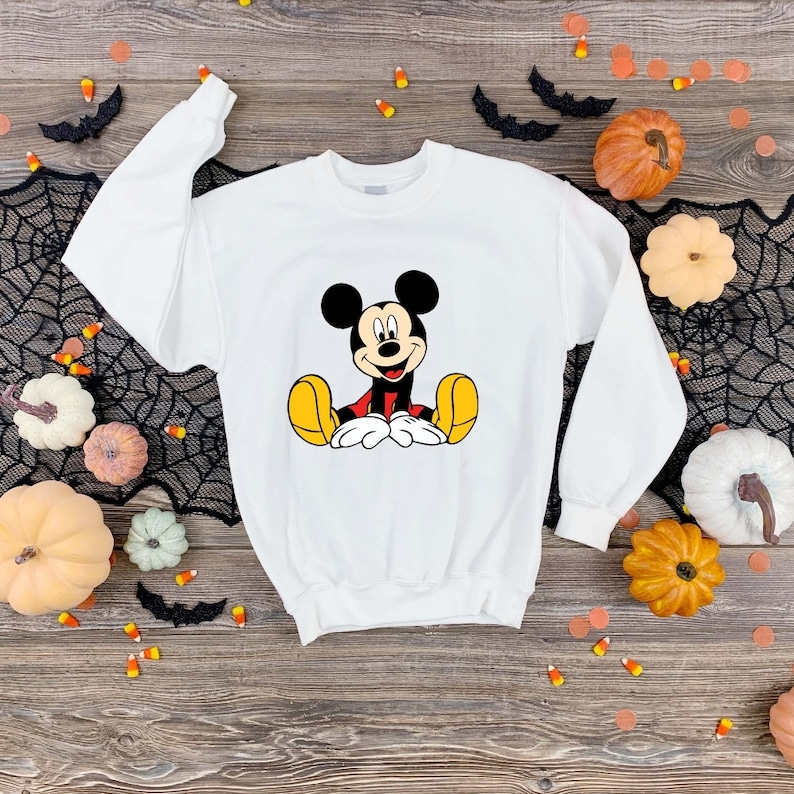 May include: White sweatshirt with a cartoon image of Mickey Mouse sitting down. The sweatshirt is on a wooden surface with pumpkins and candy corn.