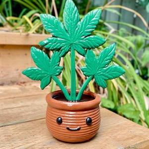 Könnte beinhalten: Ein skurriler, handgefertigter Keramiktopf mit einem lächelnden Gesicht, der drei leuchtend grüne Cannabispflanzenblätter enthält. Der Topf hat eine warme braune Farbe und steht auf einer Holzoberfläche, was einen verspielten und dekorativen Gegenstand ergibt.