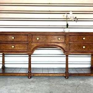 May include: A dark brown wooden console table with four drawers and a curved center section. The drawers have gold knobs. The table has four legs and a lower shelf. A white vase with flowers sits on top.