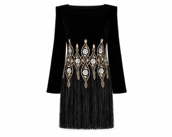 May include: A black long-sleeved dress featuring a boat neckline and structured shoulders. The waist area is adorned with an intricate gold and silver embroidered pattern of floral motifs, finished with a long black fringe hemline.