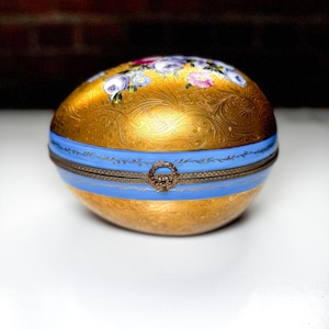 May include: A golden egg-shaped trinket box with a blue band and floral detailing. The box features a hinged lid and a decorative clasp. The top is adorned with a floral pattern in shades of purple, pink, and white.
