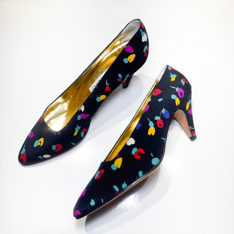 Puede incluir: Un par de zapatos de tac&oacute;n negros con un estampado floral colorido. Los zapatos tienen punta afilada, tac&oacute;n bajo e interior dorado. El estampado incluye detalles en rojo, amarillo, azul y morado.