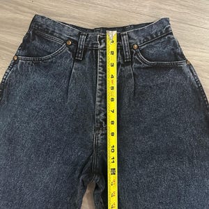 May include: Close-up of dark wash denim jeans with a high waist and pleats. A yellow measuring tape shows the length of the jeans. The jeans have brass-colored rivets and pockets.