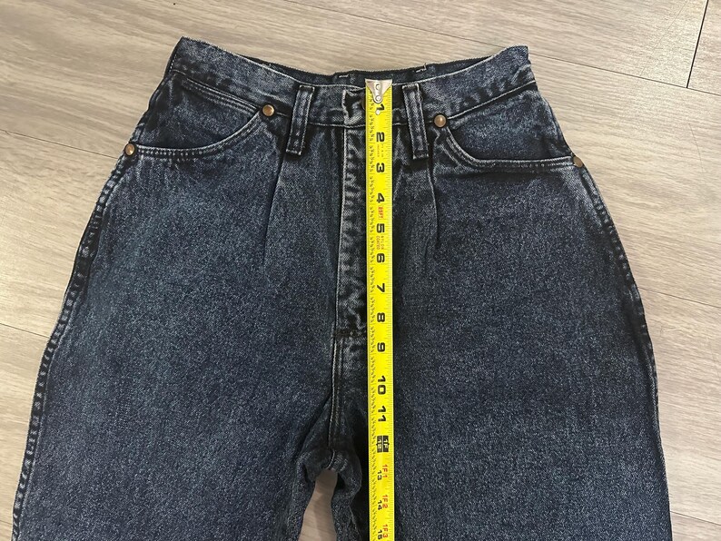May include: Close-up of dark wash denim jeans with a high waist and pleats. A yellow measuring tape shows the length of the jeans. The jeans have brass-colored rivets and pockets.