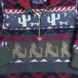 Puede incluir: Un jersey de forro polar estampado con cierre de media cremallera. El suéter presenta un diseño del suroeste con cactus, perros y formas geométricas en tonos de azul, verde, burdeos y gris. El cuello y los puños son de un verde más oscuro. La etiqueta de la marca dice "Early Winters".