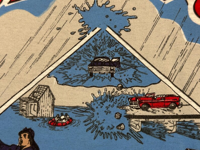 May include: A light blue t-shirt with a comic book style illustration depicting a flood scene. The image includes a house, cars, and people in a raft, with water splashing around.