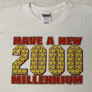 Op de afbeelding: Wit T-shirt met de tekst "HAVE A NEW 2000 MILLENNIUM" in rood en geel. Het getal 2000 is gemaakt van smiley-gezichten.