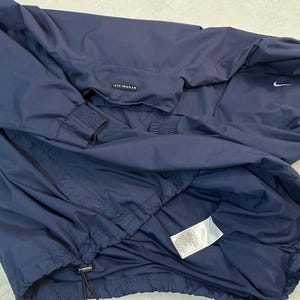 May include: Navy blue Nike Storm-FIT jacket. The jacket has a hood, a zip closure, and a drawstring at the hem. The Nike swoosh logo is visible on the upper right chest. The jacket is made of a lightweight, water-resistant material.