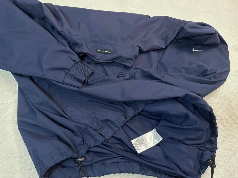 May include: Navy blue Nike Storm-FIT jacket. The jacket has a hood, a zip closure, and a drawstring at the hem. The Nike swoosh logo is visible on the upper right chest. The jacket is made of a lightweight, water-resistant material.