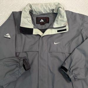 May include: A grey Nike ACG jacket featuring a high collar with a light grey interior lining, a front zipper closure, and long sleeves with elastic cuffs. The Nike swoosh logo is on the chest, and the ACG logo is on the right sleeve and inner label.