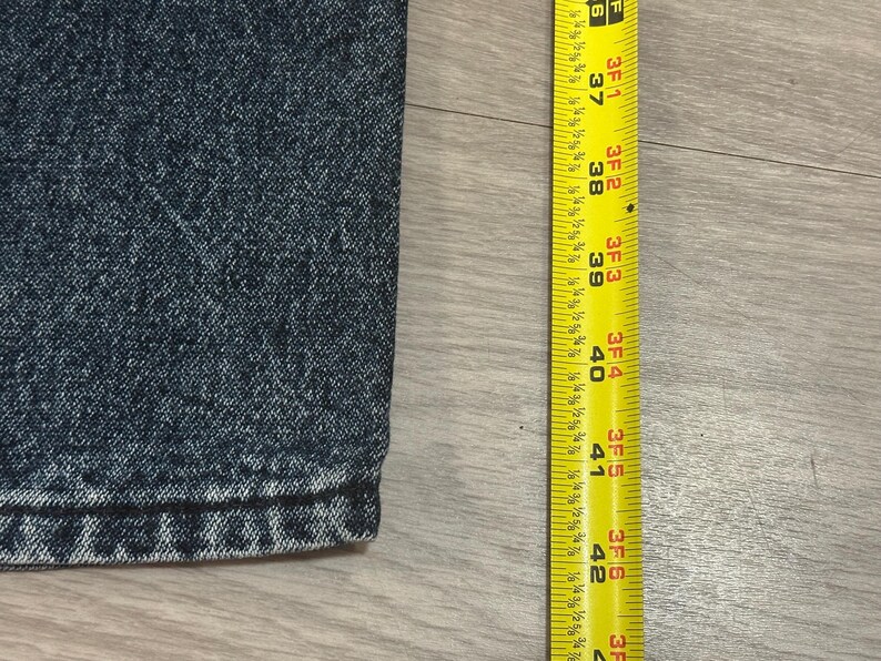 May include: Close-up of dark blue denim fabric with a yellow measuring tape. The tape shows measurements from 37 to 42 inches. The denim has a textured appearance.
