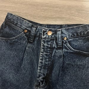 May include: Close-up of a pair of dark wash denim jeans. The jeans have a button and zipper closure, front pockets, and copper-colored rivets. The denim has a slightly faded appearance.