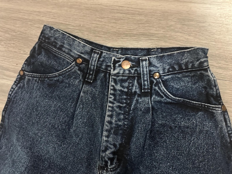 May include: Close-up of a pair of dark wash denim jeans. The jeans have a button and zipper closure, front pockets, and copper-colored rivets. The denim has a slightly faded appearance.