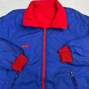 Puede incluir: Chaqueta reversible azul y roja con cierre de cremallera completo. La chaqueta tiene cuello y puños rojos, y un pequeño parche con el logotipo en el pecho. Fabricada con un material ligero.