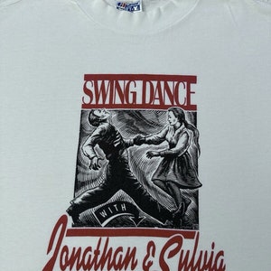 Puede incluir: Camiseta blanca con un gráfico rojo y negro de una pareja bailando swing. El gráfico dice "Swing Dance with Jonathan & Sylvia" y "© 1990 Santa Barbara Swing Dance Club".