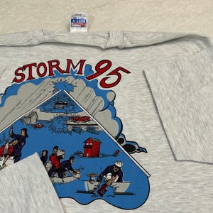 May include: A light gray long-sleeve t-shirt featuring a graphic with the text "STORM 95". The graphic depicts a storm scene with people, vehicles, and buildings. The shirt has a crew neck and long sleeves.