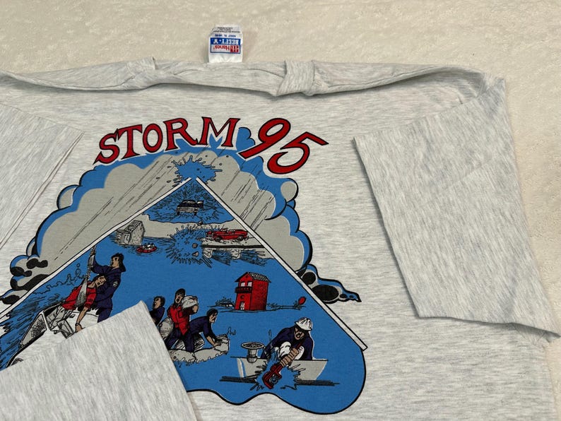 May include: A light gray long-sleeve t-shirt featuring a graphic with the text "STORM 95". The graphic depicts a storm scene with people, vehicles, and buildings. The shirt has a crew neck and long sleeves.