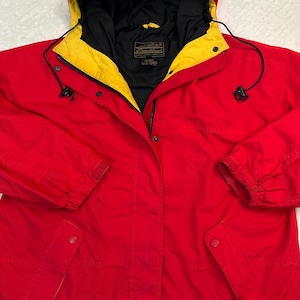 Puede incluir: Chaqueta roja con capucha, interior negro y ribete amarillo alrededor de la capucha. La chaqueta tiene cierre de cremallera y dos bolsillos delanteros. Las mangas tienen puños ajustables. La chaqueta es de material resistente.
