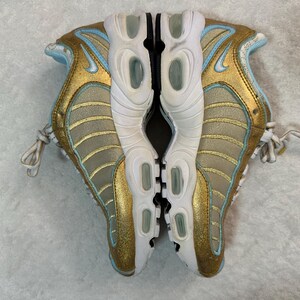 Pu&ograve; includere: Paio di scarpe sportive oro e azzurro chiaro. Le scarpe hanno suola e lacci bianchi. Il logo Nike &egrave; visibile sul lato di ogni scarpa. Le scarpe sono mostrate da una prospettiva dall'alto.