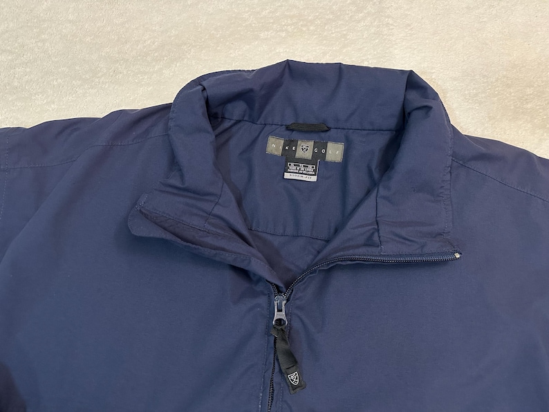 May include: A navy blue jacket with a full zip and a stand-up collar. The jacket has a tag with text and a small logo. The jacket is made of a smooth, possibly water-resistant material.