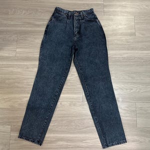May include: A pair of high-waisted, acid-wash denim jeans. The jeans have a classic five-pocket design, a button and zipper closure, and a straight leg. The denim is a dark blue color with a mottled, faded appearance. The jeans are laid flat on a light-colored wooden surface.