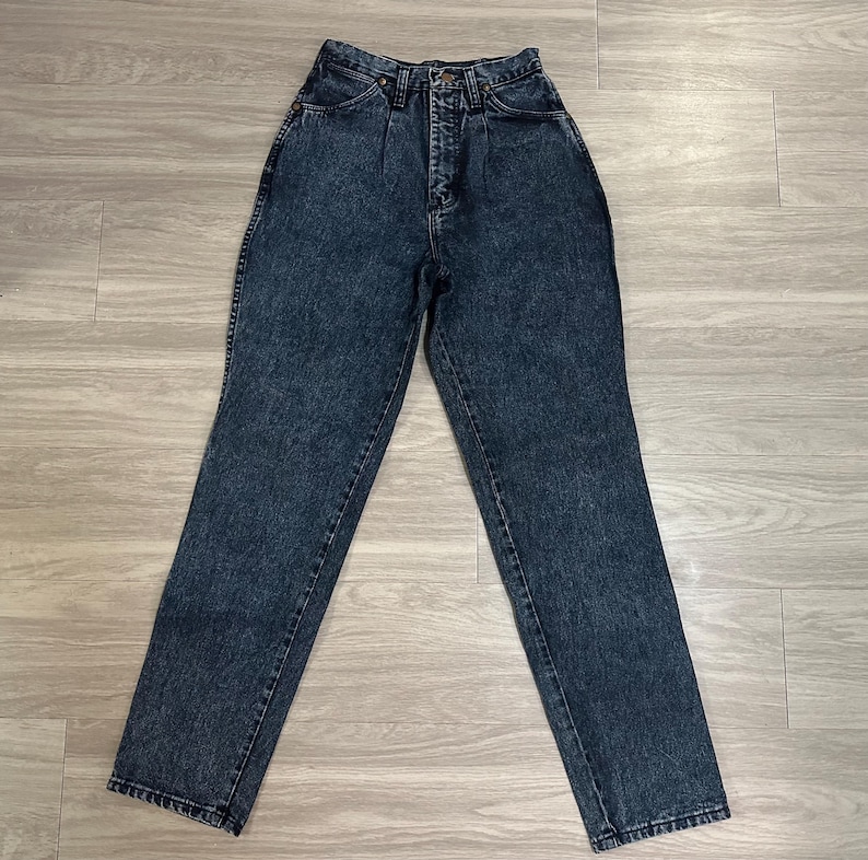 May include: A pair of high-waisted, acid-wash denim jeans. The jeans have a classic five-pocket design, a button and zipper closure, and a straight leg. The denim is a dark blue color with a mottled, faded appearance. The jeans are laid flat on a light-colored wooden surface.