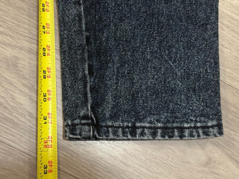 May include: Close-up of dark wash denim jeans with a yellow measuring tape. The tape shows measurements from 26 to 33 inches. The jeans have a classic five-pocket design.