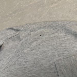 May include: Close-up of a light gray t-shirt. The shirt has a crew neck and short sleeves. The Hanes tag is visible on the left side of the shirt. The fabric has a heathered appearance.