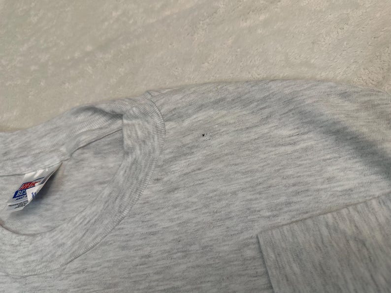 May include: Close-up of a light gray t-shirt. The shirt has a crew neck and short sleeves. The Hanes tag is visible on the left side of the shirt. The fabric has a heathered appearance.