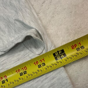 May include: A light gray fabric is measured with a yellow tape measure. The tape measure shows measurements in inches and fractions of an inch. The fabric appears to be a light-colored knit.