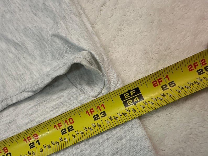 May include: A light gray fabric is measured with a yellow tape measure. The tape measure shows measurements in inches and fractions of an inch. The fabric appears to be a light-colored knit.