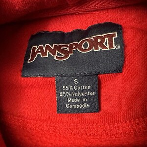 Puede incluir: Sudadera roja Jansport con una etiqueta negra que dice "Jansport" y una etiqueta m&aacute;s peque&ntilde;a que dice "S 55% Algod&oacute;n 45% Poli&eacute;ster Hecho en Camboya".