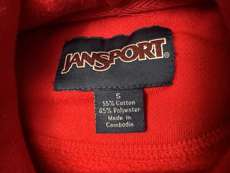 Puede incluir: Sudadera roja Jansport con una etiqueta negra que dice "Jansport" y una etiqueta m&aacute;s peque&ntilde;a que dice "S 55% Algod&oacute;n 45% Poli&eacute;ster Hecho en Camboya".