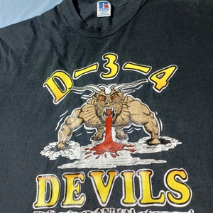 May include: Black t-shirt with a yellow and white graphic of a cartoon devil with horns and red blood dripping from its mouth. The text "D-3-4 DEVILS" is in yellow and white with the tagline "We'll make an ANIMAL of anyone!" in white.