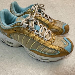 Pu&ograve; includere: Paio di scarpe da ginnastica oro e azzurro chiaro con suole e lacci bianchi. Le scarpe hanno una finitura oro metallizzato sulla tomaia, con accenti azzurro chiaro sul tallone e sulla punta. Le suole sono bianche con dettagli neri.