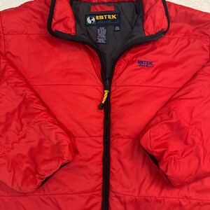 Puede incluir: Chaqueta acolchada roja brillante con cuello y cremallera negros. La chaqueta tiene el nombre de la marca "EBTEK" en azul en el pecho y un logotipo en el cuello. La chaqueta es de estilo abullonado, adecuada para climas fríos.