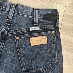 May include: Close-up of the back of dark wash Wrangler jeans. The jeans feature a leather patch with the Wrangler logo on the back pocket, along with the Wrangler label above the pocket. The jeans have copper-colored rivets and a classic denim design.