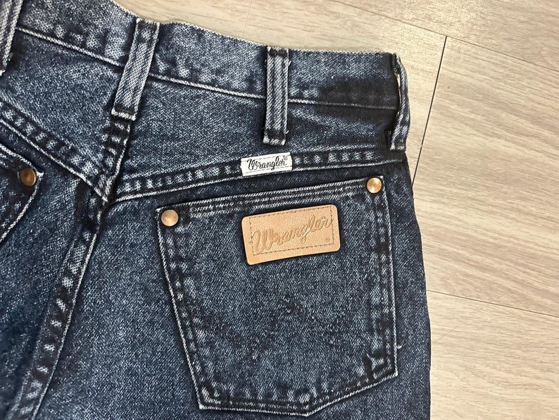 May include: Close-up of the back of dark wash Wrangler jeans. The jeans feature a leather patch with the Wrangler logo on the back pocket, along with the Wrangler label above the pocket. The jeans have copper-colored rivets and a classic denim design.