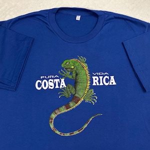 Op de afbeelding: Een koningsblauw t-shirt met korte mouwen en een afbeelding van een groene leguaan in het midden op de borst. De tekst "PURA VIDA COSTA RICA" is in witte hoofdletters gedrukt rond de illustratie van de leguaan, die een lange, gebogen staart heeft.