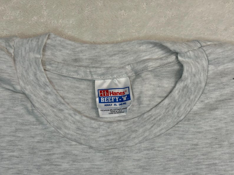 May include: Close-up of a heather gray Hanes Beefy-T adult XL t-shirt. The tag is visible, showing the brand logo and size information. The shirt is made of a soft, comfortable material.