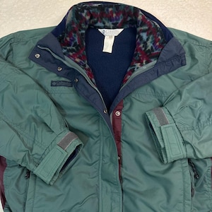 Op de afbeelding: Een teal Columbia jas met marineblauwe details. De jas heeft een gevoerde fleece voering, een ritssluiting en verstelbare manchetten. Het Columbia-logo is zichtbaar. Een vintage outdoor jas.