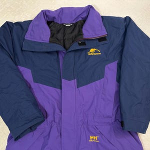 May include: A Helly Hansen jacket featuring a color-blocked design in dark blue and purple. The jacket has a high collar, a front zipper closure with a storm flap, and long sleeves. The brand logo is embroidered in yellow on the chest and lower front.