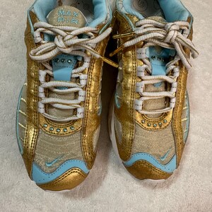 Pu&ograve; includere: Paio di scarpe sportive oro e azzurro chiaro con lacci bianchi. Le scarpe hanno un design a rete con dettagli dorati e interni azzurro chiaro. La linguetta della scarpa riporta la scritta "MAX AIR".