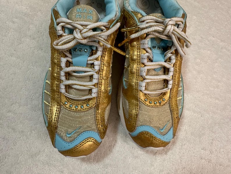 Pu&ograve; includere: Paio di scarpe sportive oro e azzurro chiaro con lacci bianchi. Le scarpe hanno un design a rete con dettagli dorati e interni azzurro chiaro. La linguetta della scarpa riporta la scritta "MAX AIR".