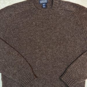 May include: A brown Lands' End crew neck sweater. The sweater features a ribbed collar, cuffs, and hem. The Lands' End label is visible at the back of the neck. The sweater is made of a textured knit fabric.