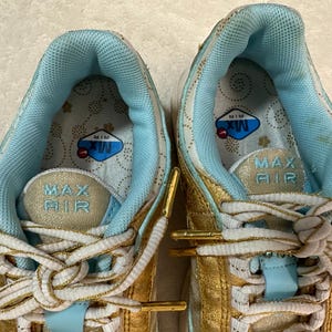 Pu&ograve; includere: Primo piano di un paio di scarpe da ginnastica oro e azzurre. Le scarpe da ginnastica hanno lacci bianchi e le parole "MAX AIR" sono visibili sulla linguetta. L'interno della scarpa &egrave; azzurro con un motivo floreale.