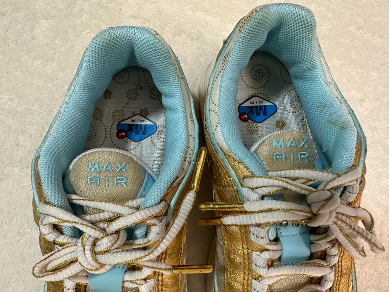 Pu&ograve; includere: Primo piano di un paio di scarpe da ginnastica oro e azzurre. Le scarpe da ginnastica hanno lacci bianchi e le parole "MAX AIR" sono visibili sulla linguetta. L'interno della scarpa &egrave; azzurro con un motivo floreale.