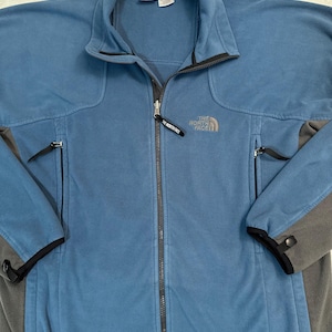 Puede incluir: Chaqueta polar azul con cremallera completa y cuello alzado. La chaqueta tiene detalles grises en las mangas y hombros, con el logotipo de la marca en el pecho. Dos bolsillos con cremallera y un tirador negro.