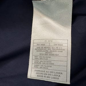 May include: A close-up of a white label with black text, attached to a navy blue fabric. The label includes the text "F1 SFD", "RN 58023", and "CA# 05553". It also lists the manufacturer's address and material composition.